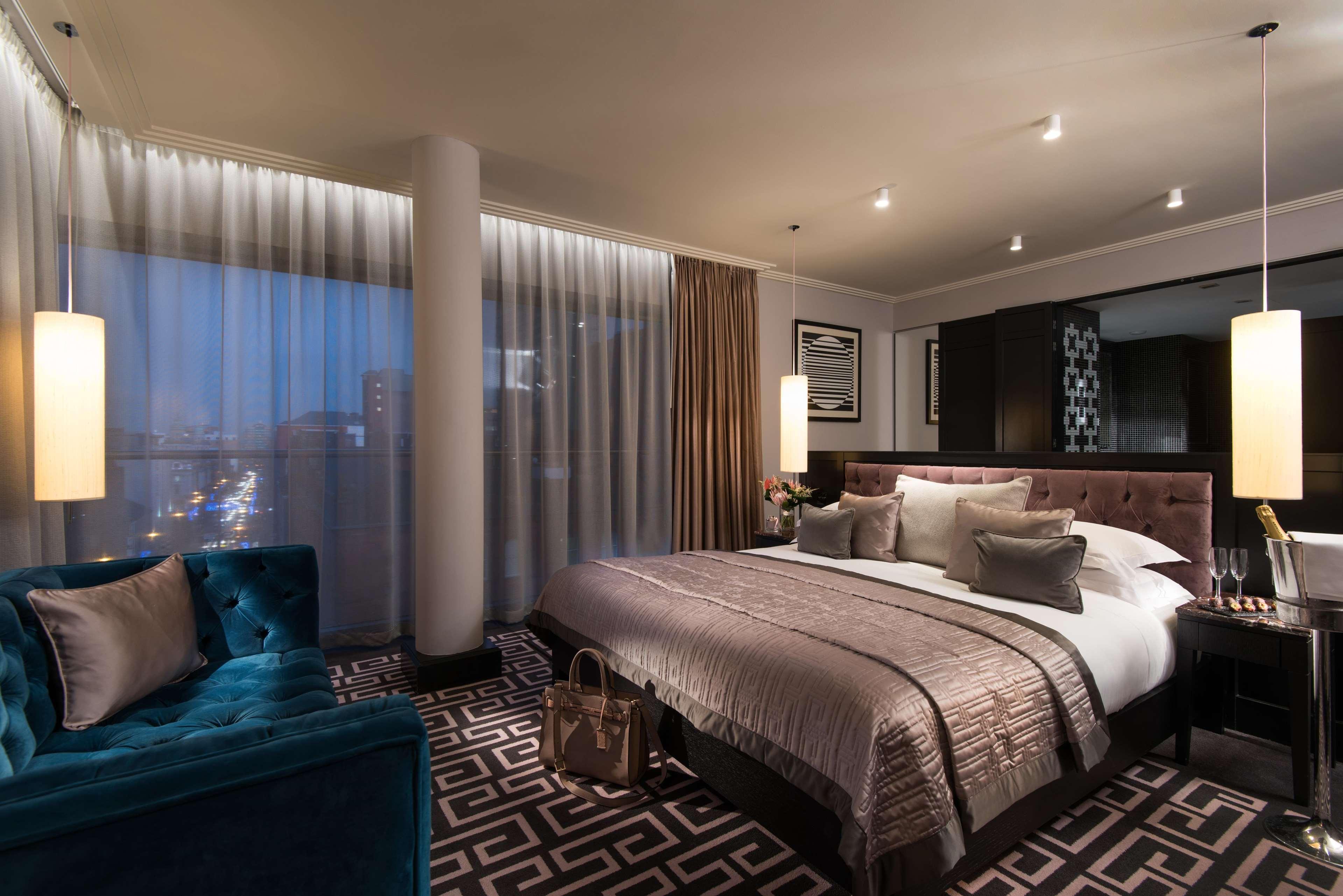The Fitzwilliam Hotel Belfast Ngoại thất bức ảnh The photo shows a luxurious hotel room with a modern design. It features a large bed with decorative pillows and a stylish bedspread, complemented by bedside lamps. To one side, there is a plush turquoise sofa and a coffee table. The room has floor-t