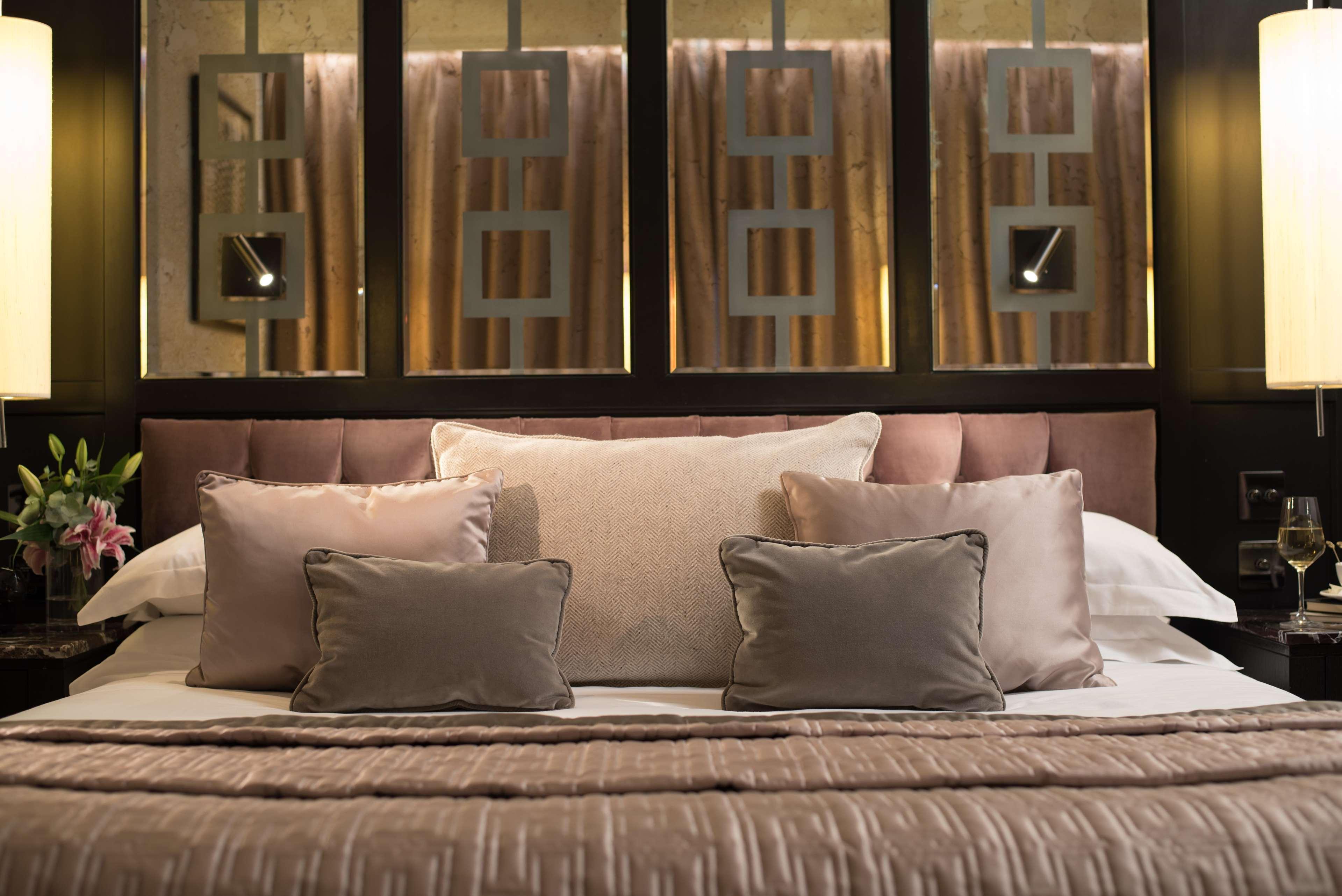 The Fitzwilliam Hotel Belfast Ngoại thất bức ảnh The photo shows a beautifully designed bed in a stylish bedroom. The bed is adorned with a soft, plush headboard and layered with various decorative pillows in shades of beige and gray. The bedding appears luxurious, with a textured blanket. Behind t