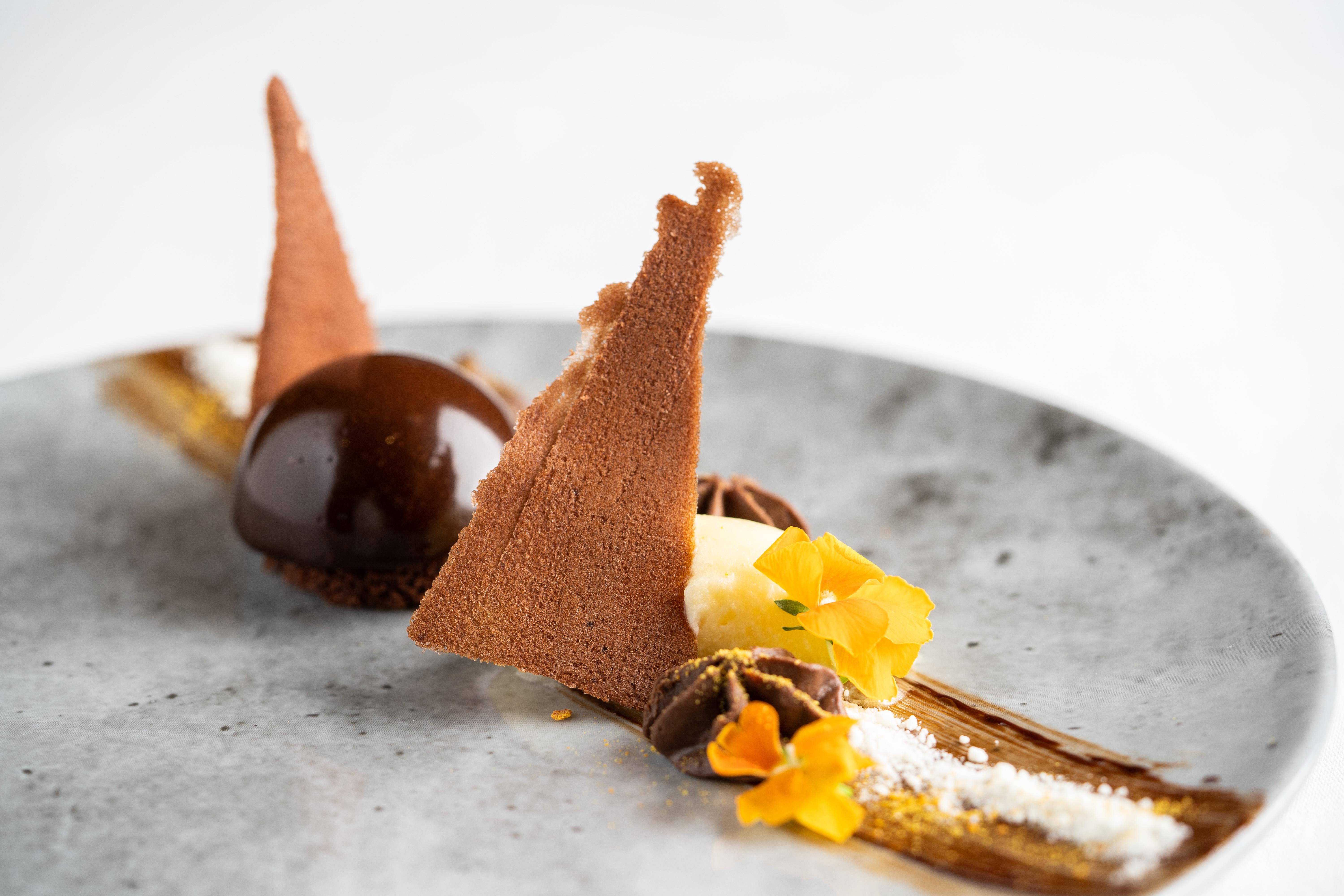 The Fitzwilliam Hotel Belfast Ngoại thất bức ảnh The photo displays an artfully plated dessert. It features a glossy chocolate sphere, accompanied by various chocolate components, including triangular chocolate tuile pieces that resemble sails. The plate is adorned with delicate garnishes such as y